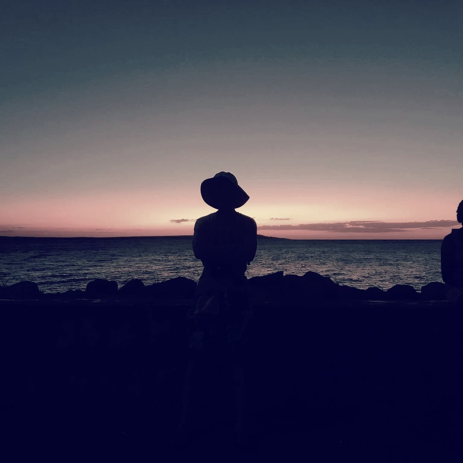 Silhouette d'une personne coiffée d'un chapeau, face à la mer au crépuscule